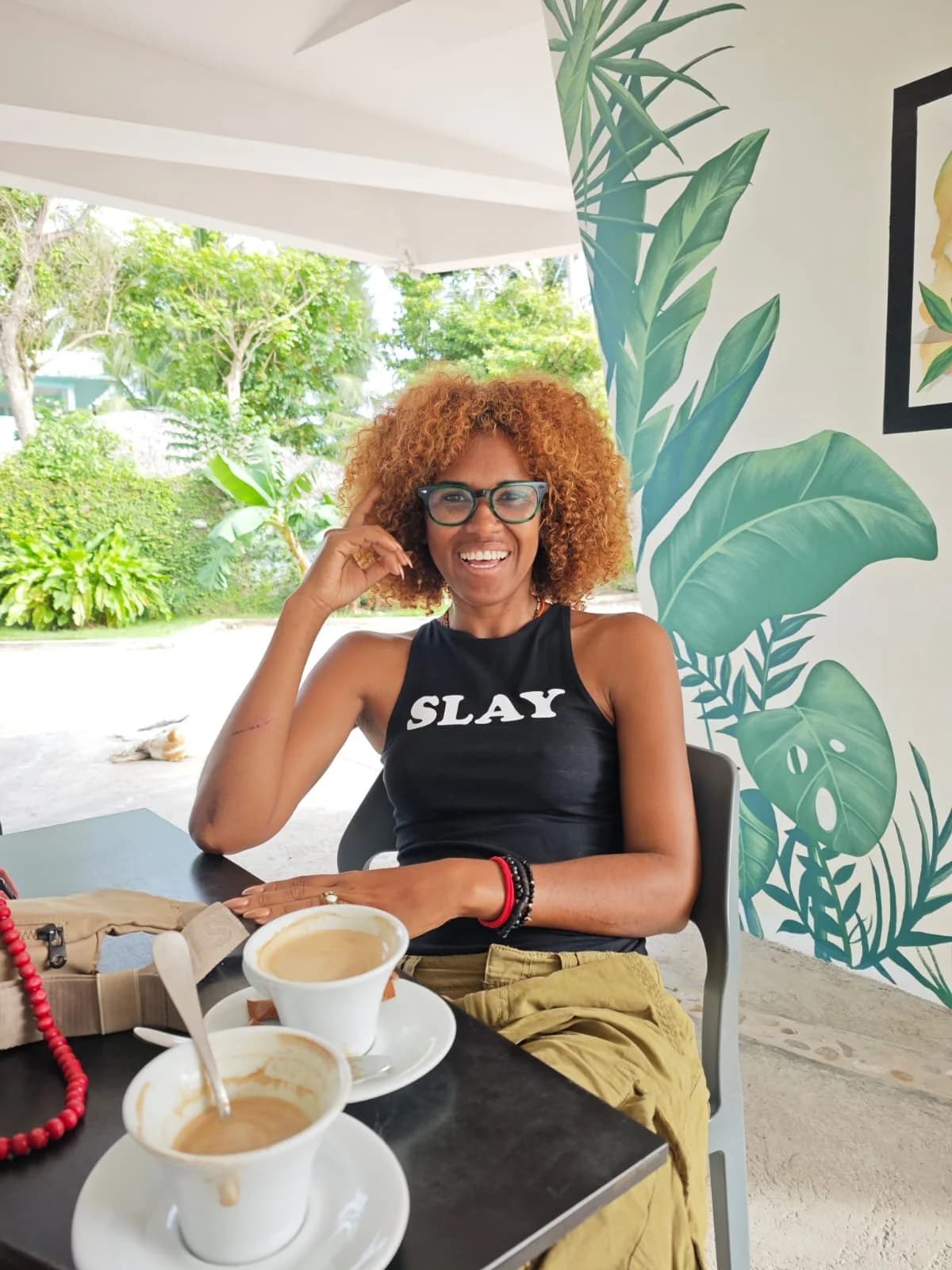 Sonriendo en la mesa de la cafetería de Dulce Vida, con dos cafés recién servidos y un mural de hojas tropicales al fondo